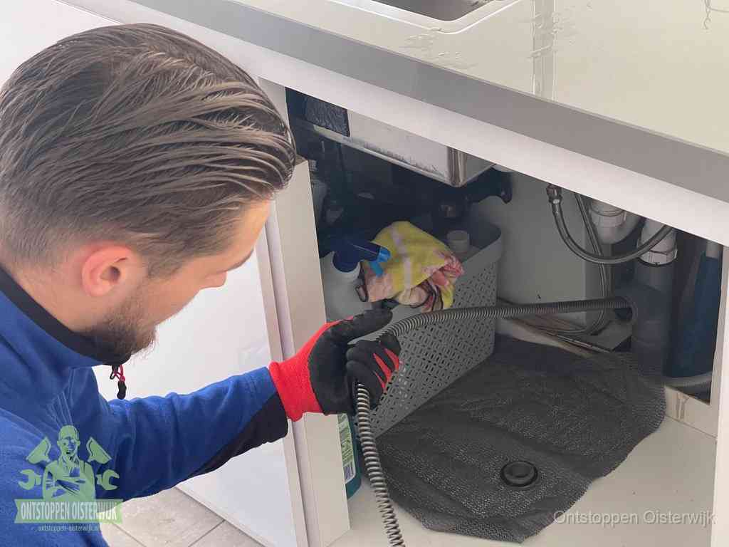Een monteur voert een flexibele ontstoppingsveer in onder het keukenkastje, langs de sifon, om de gootsteen te ontstoppen in Oisterwijk