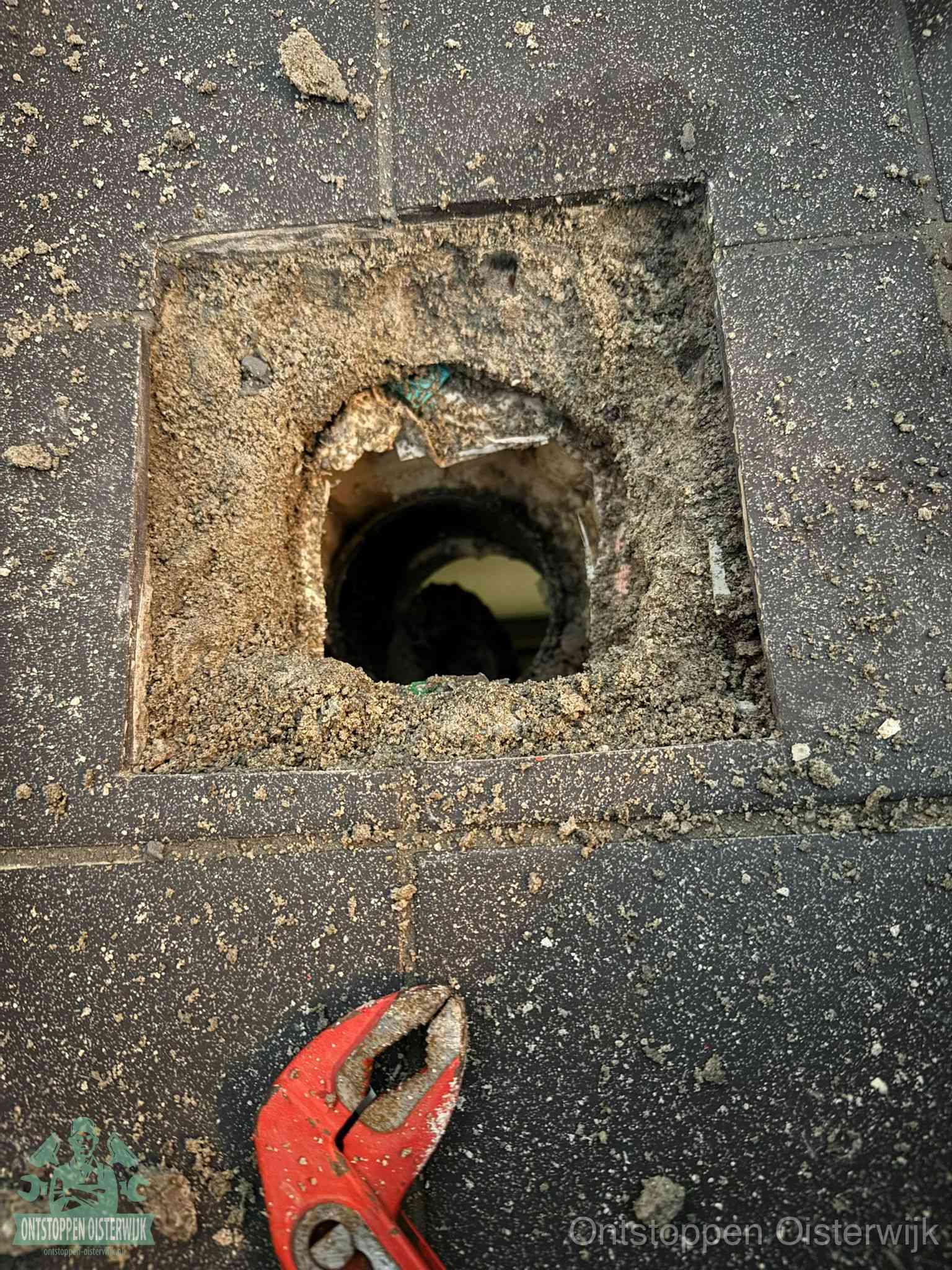 Een geopend rioolputje in een betontegelvloer met losgemaakt rooster en een zichtbare ronde afvoerbuis, het achterhalen waar de afvoerverstopping zit in Oisterwijk