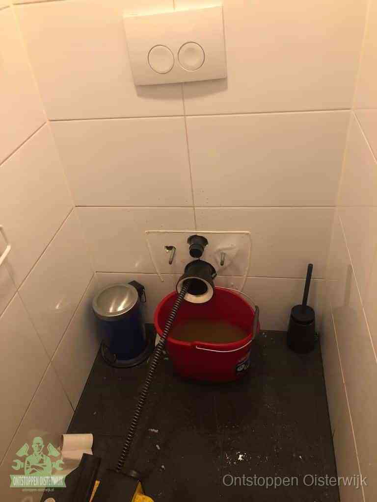 Een ontstopte wand-wc met verwijderde toiletpot waar een ontstoppingsveer in de wanduitgang verdwijnt, het achterhalen waar de verstopping zit in Oisterwijk
