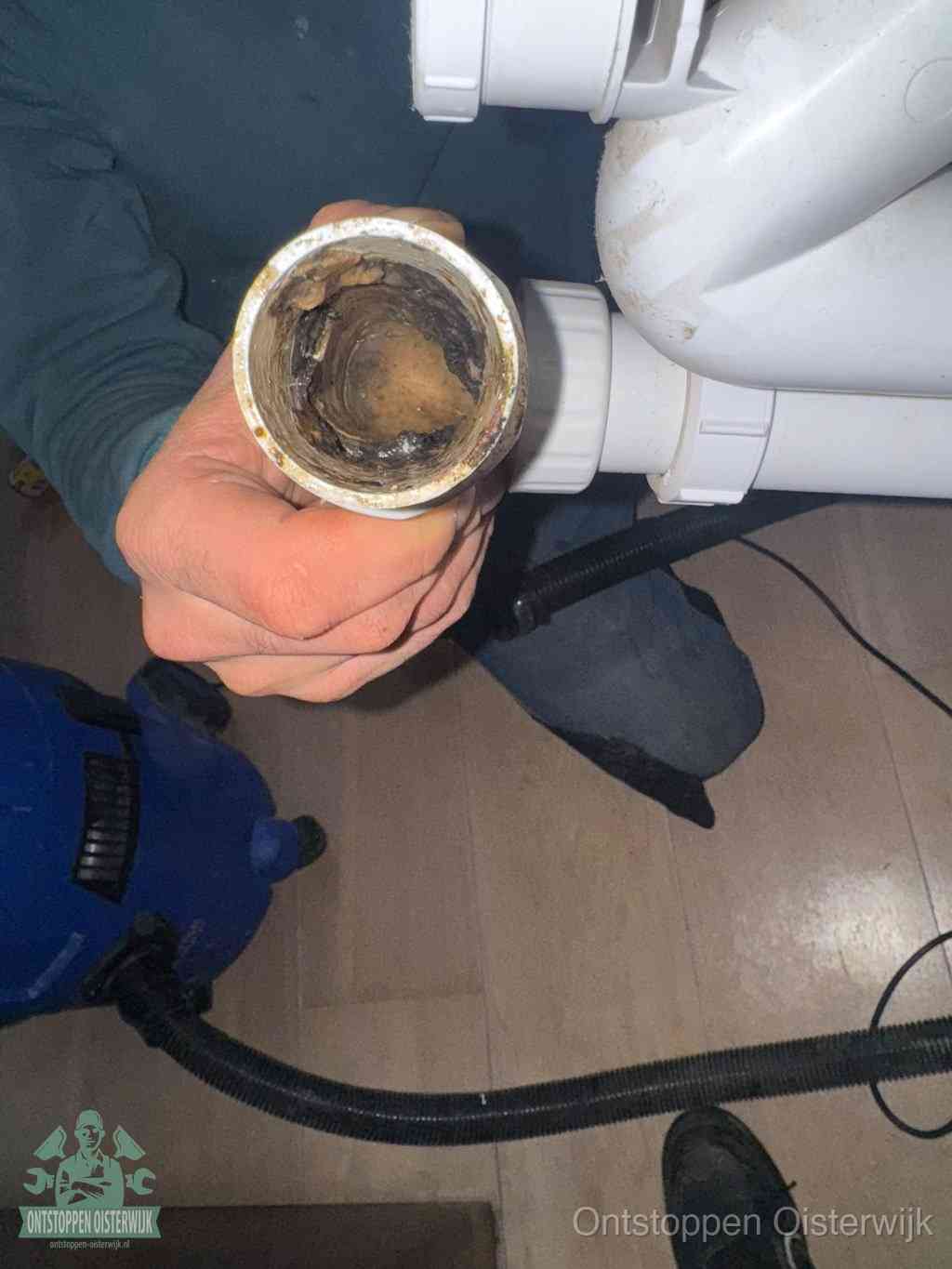 Een hand houdt een losgekoppeld PVC-afvoerbuisstuk vast dat van binnen volledig is dichtgeslibd met vet, vuil en aanslag, de meest voorkomende oorzaak van een verstopte afvoer in Oisterwijk