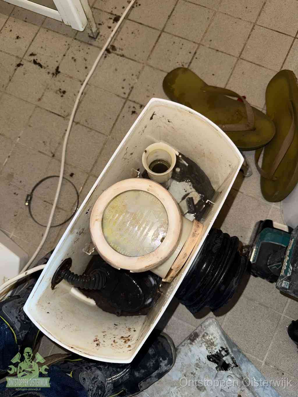 Een gedemonteerde toiletpot op de vloer met ontstoppingsveer en gereedschap ernaast, het verwijderen van een vastzittend voorwerp uit de wc in Oisterwijk